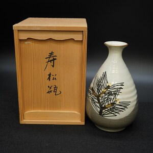Puede incluir: Una botella de sake de cerámica con un diseño de rama de pino y una caja de madera. La botella es de color blanquecino con una ilustración marrón y negra. La caja de madera tiene caracteres japoneses.