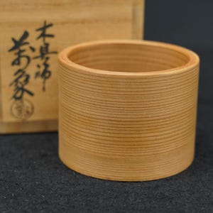 以下が含まれることがあります： 水平な木目が目立つ円筒形の明るい色の木製カップ。カップの後ろには、黒い日本語の書体と円形のスタンプが押された明るい色の木箱が置かれています。