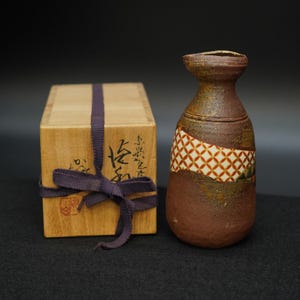 以下が含まれることがあります： 幾何学模様の茶色の陶器製酒瓶と、紫色のリボンで結ばれた木箱。 瓶は独特の形状と質感で、伝統的な職人技を思わせます。 木箱には日本語の文字が書かれています。