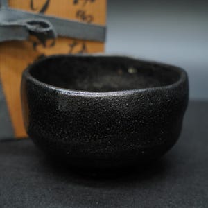 Könnte beinhalten: Eine kleine, schwarze Keramikschale mit strukturierter Oberfläche. Die Schale hat eine abgerundete Form und eine dunkle, matte Oberfläche. Eine Holzkiste mit japanischem Text und einem grauen Band befindet sich im Hintergrund.
