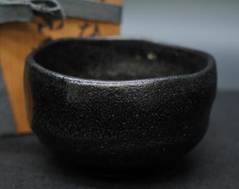 May include: A small, black ceramic bowl with a textured surface. The bowl has a rounded shape and a dark, matte finish. A wooden box with Japanese text and a gray ribbon are in the background.