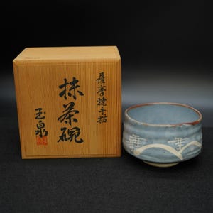 Może przedstawiać: Niebieska ceramiczna miska do matchy z białym wzorem fal i drewnianym pudełkiem. Pudełko ma japoński tekst i czerwoną pieczęć. Miska ma brązową krawędź i jasnoniebieskie wnętrze.