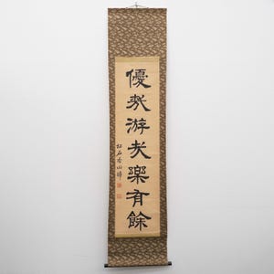 Op de afbeelding: Een verticale Japanse rol met zwarte kalligrafie op een crèmekleurige achtergrond. De boven- en onderkant hebben een bruine stof met patroon. De rol is opgehangen aan een metalen staaf.
