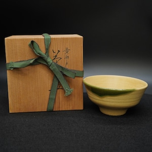 Puede incluir: Un cuenco de cerámica amarillo claro con un borde verde se encuentra junto a una caja de madera atada con una cinta verde. El cuenco tiene forma redondeada y base con pie. La caja tiene caracteres japoneses.