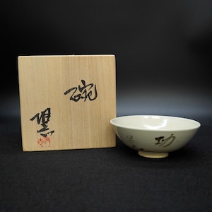 Puede incluir: Un cuenco de cerámica color crema con forma redondeada y un pequeño pie. El cuenco tiene caligrafía negra en el lateral. A la izquierda del cuenco hay una caja de madera con caligrafía negra y sellos rojos.