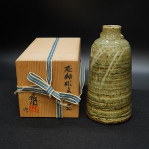 May include: A handcrafted, green and brown ceramic vase with a textured surface, next to a wooden box tied with a blue and white ribbon. The box has Japanese text and a red stamp.