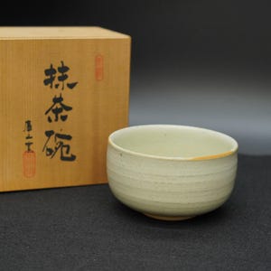 Puede incluir: Un cuenco de matcha de cerámica verde claro con una superficie texturizada y un borde ligeramente ensanchado. El cuenco está junto a una caja de madera con caracteres japoneses impresos. El cuenco tiene un diámetro de aproximadamente 11,4 cm.