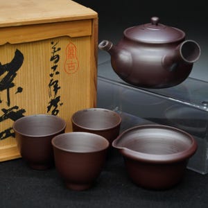 以下が含まれることがあります： 茶器セット。茶色の陶器製のティーポット、カップ4つ、サービング皿。木箱には日本語の文字が書かれています。ティーポットにはサイドハンドルと蓋が付いています。