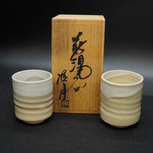 以下が含まれることがあります： 淡いベージュと白の配色で、リブのデザインが施された2つのセラミックティーカップ。日本の書道が施された木箱と一緒に展示されています。カップは素朴で手作りの外観をしています。