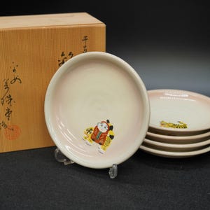 以下が含まれることがあります： 淡いピンクとクリーム色のグラデーションが施された日本の陶器のプレートのセット。各プレートには、伝統的な人物のカラフルなイラストが描かれています。背景には、日本語の文字が入った木箱があります。