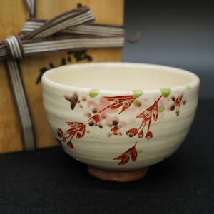 May include: Cream-coloured ceramic tea bowl with a floral design in red, pink, and green. The bowl has a round shape and sits on a small, reddish-brown base. A wooden box with a ribbon is in the background.