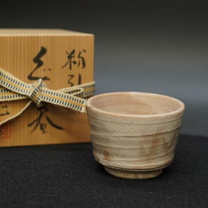 May include: A handmade, beige ceramic tea cup with a textured, ridged exterior. The cup sits on a small base and is next to a wooden box tied with a ribbon. The box has Japanese characters on it.
