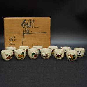May include: A set of twelve small, off-white ceramic cups with hand-painted animal designs, displayed in front of a wooden box with Japanese calligraphy. The cups feature various animals in vibrant colors, including a dog, a tiger, and a cow.