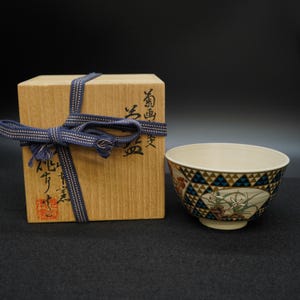 以下が含まれることがあります： クリーム色の内側と、青と金の三角形の幾何学模様と花の模様が特徴の装飾的な外側を備えたセラミックボウル。 ボウルの隣には、青いリボンで結ばれた木箱があります。