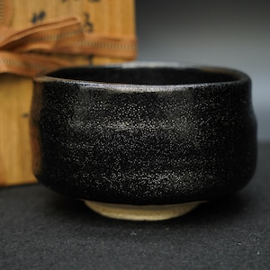 May include: A black ceramic tea bowl with a textured, speckled surface and a smooth, rounded rim. The bowl features a wide, deep basin and sits on a light beige, unglazed circular foot. The exterior finish has a subtle, granular appearance.