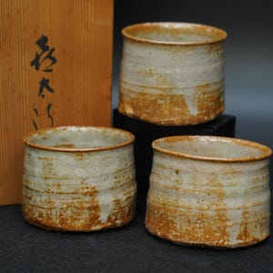 以下が含まれることがあります： 土の質感を感じさせる茶褐色の釉薬とオフホワイトの釉薬が混ざり合った、円筒形の陶器のカップ3点。表面にはわずかな凹凸と横方向の筋があり、口が広く開いた素朴なデザインが特徴的です。
