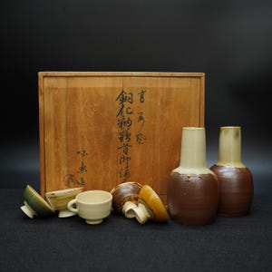 May include: A Japanese sake set with a wooden box, two brown and cream-colored bottles, and several small cups. The box has Japanese characters on it. The cups and bottles are arranged on a black surface.