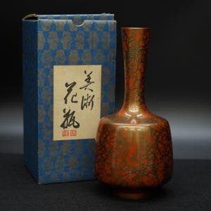 Puede incluir: Un jarrón decorativo con un patrón craquelado rojo y dorado, acompañado de una caja azul con un diseño a cuadros y texto japonés. El jarrón tiene un cuello delgado y una base redondeada.