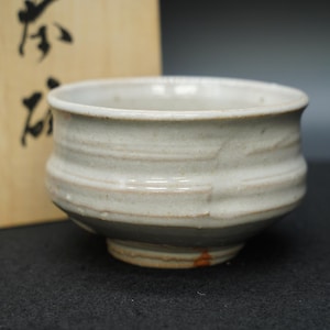 Puede incluir: Un cuenco de cerámica blanco roto hecho a mano con un diseño ancho y poco profundo. El cuenco presenta estrías horizontales y un borde ligeramente ensanchado. La base tiene una pequeña zona sin esmaltar con un toque de naranja. El cuenco es probablemente para servir comida o como pieza decorativa.