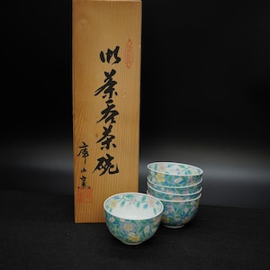 以下が含まれることがあります： 木箱に入った、花柄のティーボウル4個セット。ボウルは、青、黄色、白の花柄模様のティール色の背景が特徴です。木箱には日本語の文字と赤いスタンプが施されています。