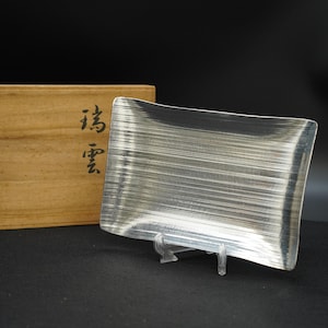 以下が含まれることがあります： ブラシ仕上げの長方形の銀色の金属トレイ。透明なスタンドに展示されています。背景には、日本語の文字が入った木箱が部分的に見えます。トレイはわずかに湾曲した形状をしています。