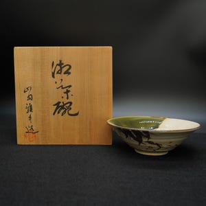 Könnte beinhalten: Keramikschale mit grünem und weiß glasiertem Rand und dekorativem Blättermuster. Die Schale steht neben einer Holzkiste mit japanischer Kalligraphie. Die Kiste hat einen roten Stempel und mehr japanischen Text.