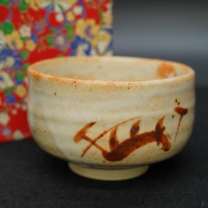 以下が含まれることがあります： クリーム色のセラミックティーボウル。茶色の抽象的なデザインが施されています。ボウルは幅が広く丸みを帯びた形状で、わずかにテクスチャ加工された表面です。背景には、赤と花の模様の箱があります。