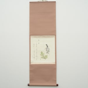 Puede incluir: Una pintura japonesa antigua en rollo, de forma rectangular vertical. El rollo es de color rosa apagado, con una pintura detallada de una figura con vestimenta tradicional, caligrafía y un borde decorativo. Se cuelga con un lazo de metal dorado.