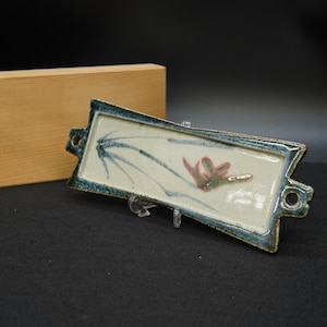 Puede incluir: Plato rectangular de cerámica con interior color crema y borde azul oscuro. El plato presenta un diseño pintado a mano de líneas azules y un motivo floral rojo y verde. El plato tiene asas en cada extremo.