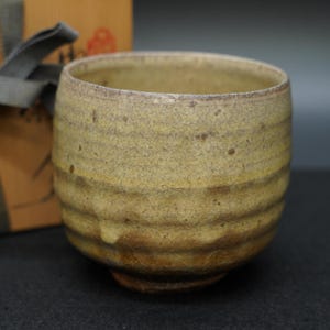 May include: A handcrafted, round ceramic tea bowl with a textured, crackled surface. The bowl features horizontal ridges and a gradient of colors, transitioning from a light yellow to a darker brown. A gift box with a gray ribbon is partially visible in the background.