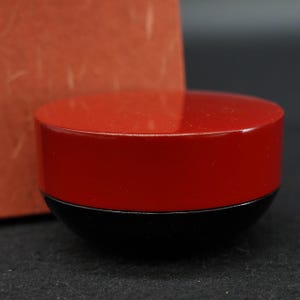 Può includere: Un piccolo contenitore rotondo con un coperchio rosso lucido e una base nera opaca. Il coperchio è piatto e circolare, si adatta saldamente al fondo a forma di ciotola. L'oggetto presenta una finitura liscia e lucida sulla parte superiore.
