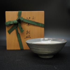 以下が含まれることがあります： 灰色の陶器のボウルに、明るい色の液体が入っています。ボウルの後ろには、緑のリボンで結ばれた木箱があります。箱には日本語の文字と数字が書かれています。ボウルは手作り感のある質感です。