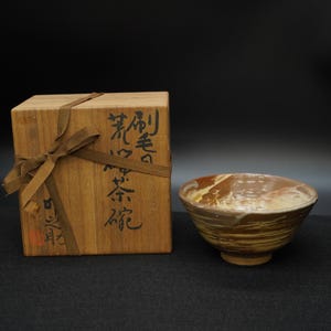 Può includere: Ciotola da tè in ceramica con un motivo marmorizzato marrone e crema. La ciotola è presentata con una scatola di legno legata con un nastro marrone, con calligrafia giapponese.
