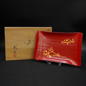 以下が含まれることがあります： 金色の幾何学模様が施された赤い長方形のサービングトレイ。トレイは透明なスタンドに展示されています。背景には、日本の書道とイラストが描かれた木箱があります。