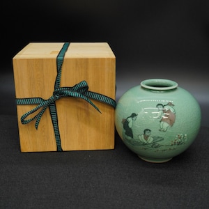 May include: A celadon green ceramic vase with a round shape and a painted scene of figures. The vase is next to a wooden box tied with a dark green ribbon. The box is made of light-colored wood.