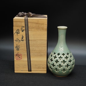 May include: A pale green ceramic vase with a cross-shaped cut-out pattern. Beside it is a wooden box tied with a brown ribbon. The box features Asian calligraphy and a red stamp.