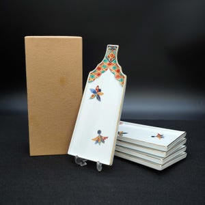 Puede incluir: Un juego de fuentes de cerámica blanca con un diseño floral decorativo. Las fuentes tienen un borde dorado y van acompañadas de una pieza de servicio a juego y una caja de cartón. La pieza de servicio tiene un patrón geométrico colorido.