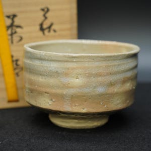 May include: A beige ceramic tea bowl with a wide, rounded shape and a footed base. The bowl has a textured surface with subtle variations in color, including light brown and white. The rim is smooth, and the bowl appears to be handmade.