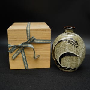 Puede incluir: Una botella de sake de cerámica con un esmalte verde claro moteado y un diseño abstracto negro. Una caja de madera con una cinta azul y blanca está al lado de la botella. La botella tiene un borde marrón oscuro.