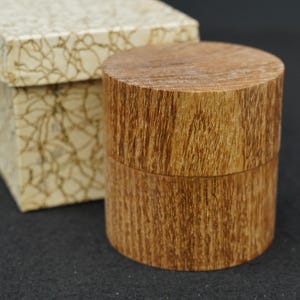 Può includere: Un contenitore cilindrico in legno con una venatura naturale visibile, dotato di un coperchio abbinato. Il contenitore è posizionato accanto a una scatola quadrata color bianco sporco con un delicato motivo a ragnatela marrone scuro.