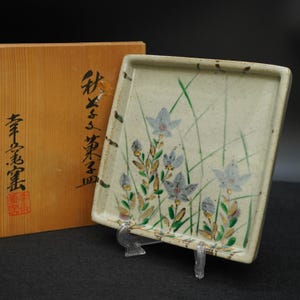 以下が含まれることがあります： 淡い青色の花と緑と金のアクセントが特徴の、長方形のセラミックプレート。透明なスタンドに飾られており、背景には木箱があります。木箱には日本語のテキストが書かれています。