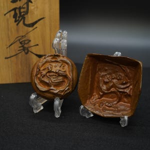 以下が含まれることがあります： 透明なスタンドに展示された、2つの茶色の彫刻された木製オブジェクト。1つは花の模様の円形、もう1つは詳細なレリーフの正方形です。背景には日本語のテキストが入った木箱があります。