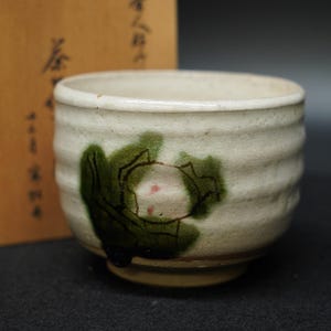 以下が含まれることがあります： クリーム色のセラミックティーボウル。手描きの緑とピンクの花柄。ボウルは、テクスチャ加工されたリブ付きの外面と丸みを帯びた形状をしています。木製の箱が背景に部分的に見えます。