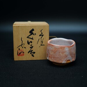 以下が含まれることがあります： 赤茶色の外観と白い内装の、手作りの小さな陶器のカップ。 カップは小さな円形のベースの上にあります。 その隣には、日本の書道が施された木箱があります。
