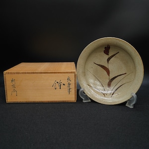 May include: A beige ceramic plate with a floral design in brown and red, displayed on a clear stand. Beside it is a wooden box with Japanese characters. The plate has a slightly raised rim.