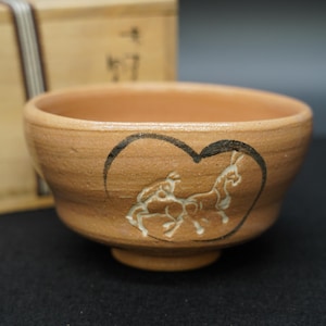 May include: A small, round, earthenware bowl with a light brown exterior. The bowl features a black heart-shaped outline with white horse figures. The bowl is likely used for serving food or drink.