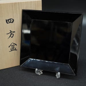 以下が含まれることがあります： 光沢のある仕上げの正方形の黒い漆塗りのトレイ。明るい色の木製収納ボックスの前に配置されています。ボックスの前面には、黒い日本語の書道文字で「四方盆」というテキストが記されています。