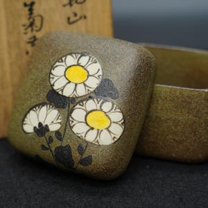 以下が含まれることがあります： 白い花と黄色の中心部、黒い茎と葉で飾られた、正方形のオリーブグリーンのセラミックボックス。箱は開いており、内部が見えます。花は白い花びらと黄色の中心部、黒い茎と葉を持っています。
