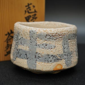以下が含まれることがあります： クリーム色のベースとグレーの幾何学模様が施された、質感のあるセラミックティーカップ。カップはわずかに不規則な形状で、底に小さなオレンジブラウンの斑点があります。背景には日本語の文字が入った木箱があります。