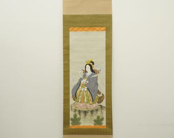 May include: A traditional Japanese scroll painting featuring a seated figure in a gray robe with gold and colorful accents. The artwork is framed with a gold border and mounted on a tan scroll with wooden hanging hardware.